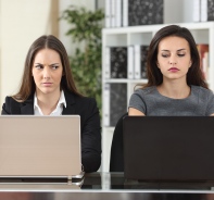 Zwei Frauen sitzen nebeneinander vor Laptops und werfen einander neidische Blicke zu.